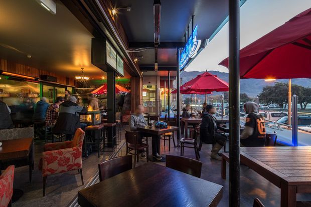 A lively commercial restaurant or bar interior opening onto an outdoor terrace. The space features dark timber tables, high stools, upholstered seating, a bar counter, warm pendant and track lighting, exposed dark ceiling elements, and large open glazing. Red patio umbrellas and outdoor seating are visible beyond, with a mountain and urban waterfront setting in the distance.