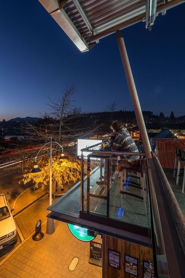 A dusk exterior view from an elevated balcony or rooftop platform overlooking a lively urban streetscape. The image shows a glass balustrade, dark metal framing, exposed roof structure and a prominent angled support post, with illuminated signage and venue frontage below. City lights, trees, distant hills and surrounding buildings form the background.