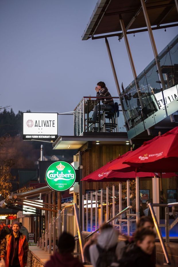Exterior evening photograph of a multi-level hospitality venue with a glass-fronted upper balcony, timber-clad lower facade, metal railings, large roof overhang, and outdoor dining terraces. Red umbrellas, illuminated signage, and patrons indicate a restaurant and bar setting. The building is positioned in a wooded or mountainous outdoor environment, though the surrounding landscape is secondary to the commercial facade.
