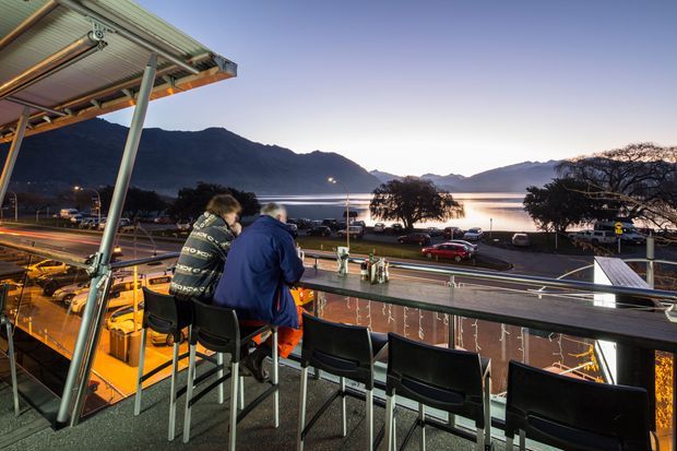 An elevated outdoor dining or viewing deck with a row of bar-height stools and tables beneath a corrugated canopy. Two people sit at the railing overlooking a road, lakeside waterfront, and silhouetted mountains at dusk. The space has exposed metal supports, dark balustrades, and warm artificial lighting.