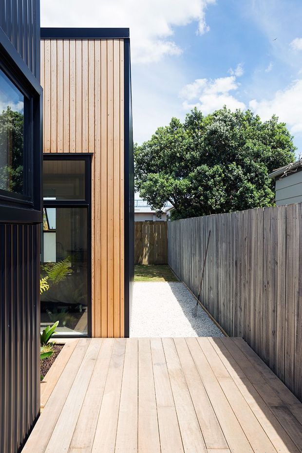 A narrow side courtyard and timber deck beside a contemporary building. The facade combines vertical light timber cladding with dark metal-framed glazing and trim. A pale timber deck leads to a white gravel path bordered by a tall weathered timber fence, with planted greenery and neighbouring structures visible beyond. Bright, sunny outdoor architectural photograph.