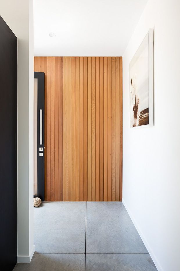 A bright minimalist entryway or hallway featuring a full-height vertical timber slat feature wall and a dark contemporary front door with a long vertical handle. Large-format pale grey floor tiles, white walls and ceiling, and a framed artwork complete the restrained interior.