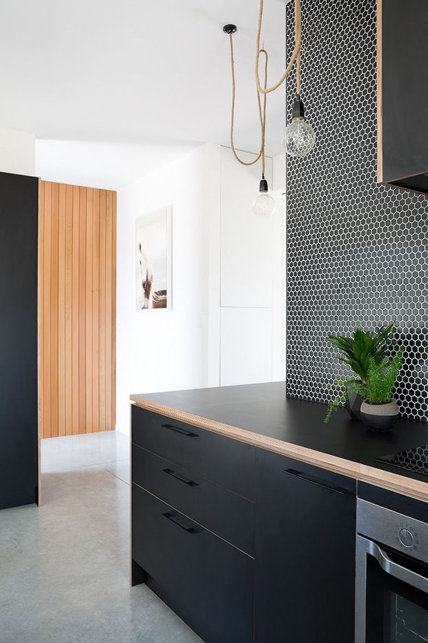 Contemporary kitchen with matte black cabinetry and benchtop, a dark hexagonal tile splashback, pale concrete-look flooring, and warm vertical timber wall panelling. Minimal pendant lights with exposed cord loops and a small potted plant add detail to the monochrome scheme.
