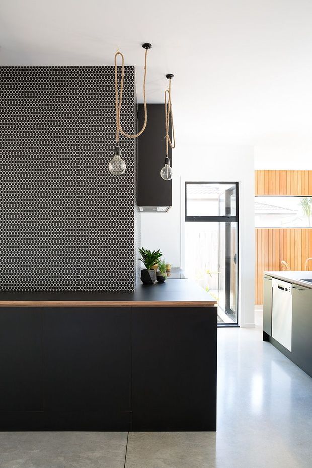 A contemporary kitchen with a large black island, dark cabinetry, a dense black-and-white penny-round tile splashback, and suspended globe pendant lights with natural cord. Pale polished concrete flooring and white walls create a bright, high-contrast interior, with timber cladding visible through the glazed doorway.