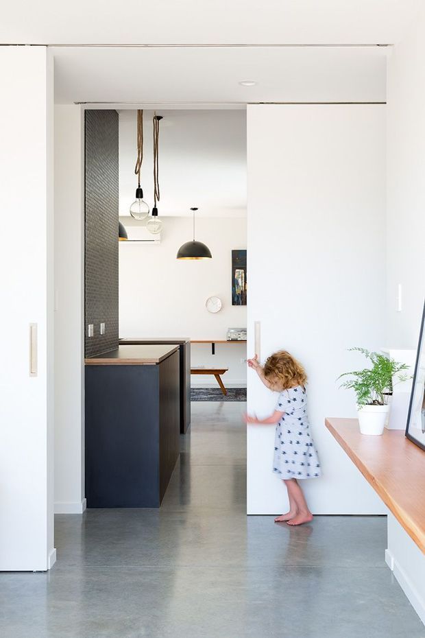 Bright minimalist interior view through a wide opening into a kitchen and dining area. The space features smooth white walls, a dark textured tile feature wall, dark kitchen island cabinetry with a timber benchtop, polished concrete flooring, and simple pendant lights. A child is pictured opening a concealed or flush white door, while a timber-topped console and potted plant sit in the foreground.