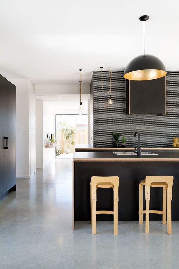 A bright, contemporary kitchen focused on a dark island with an integrated sink, black tapware, timber stools, and a large black dome pendant light. The kitchen features a small patterned geometric tile splashback, dark cabinetry, pale smooth flooring, white walls, and a view through the adjoining hallway toward a light-filled outdoor area.