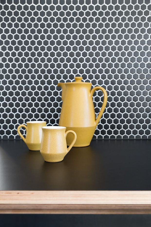 Close-up interior still life of a mustard-yellow ceramic teapot and two matching cups placed on a dark countertop. Behind them is a prominent black and white hexagonal tile backsplash, with a layered light timber edge visible at the lower portion of the image.