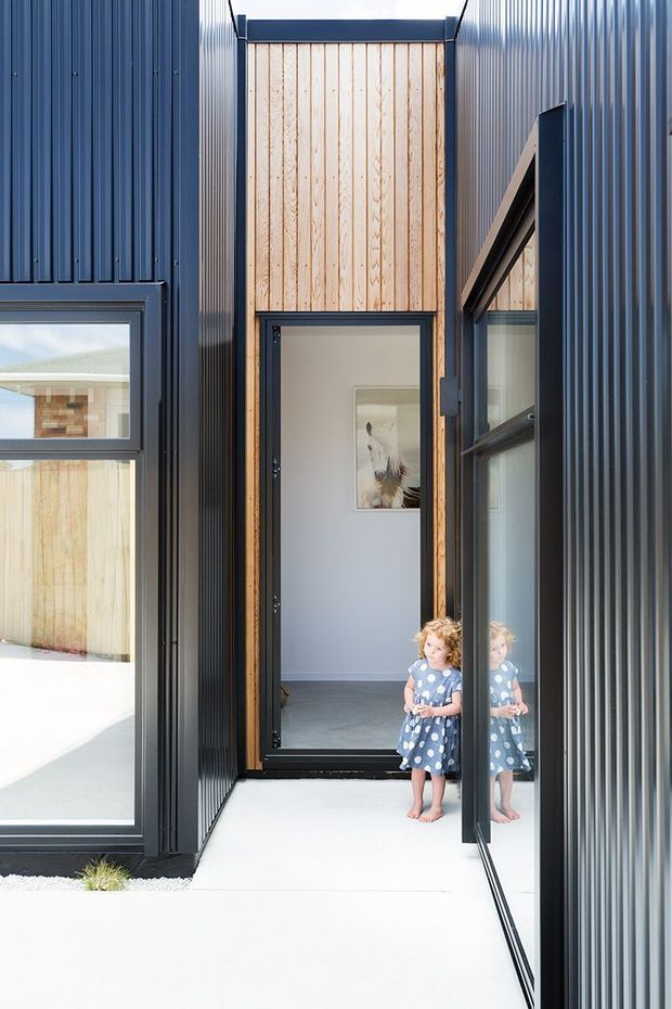A narrow, bright entry passage between dark vertically ribbed metal-clad volumes. The passage features a full-height timber-panelled wall, black-framed glazed doors and windows, a pale concrete floor, and a child visible near the entrance with a reflection in the glazing. The composition highlights a compact contemporary architectural entry and contrasting timber and dark metal materials.