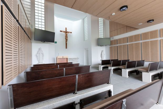 Bright contemporary chapel interior with rows of dark timber pews, pale terrazzo-like flooring, extensive light timber slatted wall panelling, white walls, glass-block clerestory windows, and wall-mounted screens. A crucifix and religious statues establish the space as a church or chapel. The ceiling is clad in light timber boards with simple circular recessed fixtures.