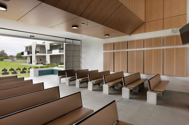 A bright, contemporary chapel or communal gathering space with rows of brown upholstered bench seating on concrete block supports. The room features a white tiled floor, a warm timber-lined angled ceiling, white walls, and extensive timber louvred wall panels. Full-height glazing opens to a landscaped courtyard and a modern building beyond. Minimal black lighting and a slim black metal screen add architectural detail.