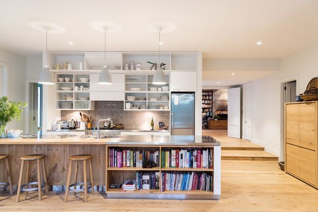 Bright open-plan kitchen with a large timber-fronted island, integrated open bookshelf, pale wood flooring, white open shelving, stainless-steel refrigerator, tiled splashback, and pendant lighting. A timber stairway and hallway lead to adjoining areas, with a tall timber storage cabinet on the right.