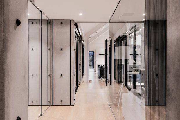 A long, refined interior corridor with pale timber flooring, exposed concrete wall panels, extensive black-framed glass partitions and doors, and a glazed ceiling or overhead glass element. The hallway opens toward a brighter living area and appears spacious, clean-lined, and contemporary with strong reflective surfaces.