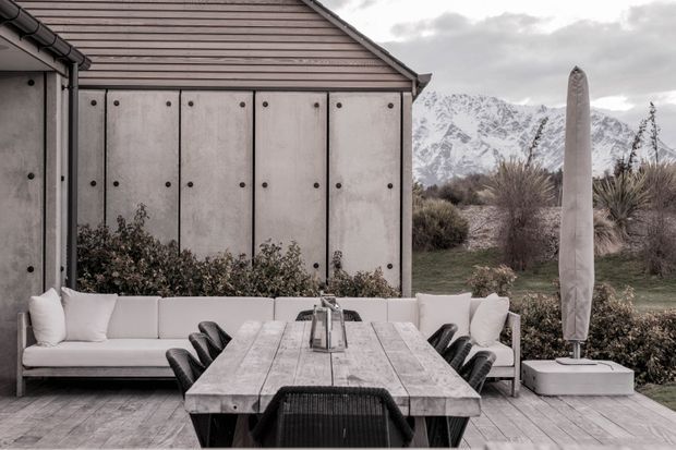 Outdoor dining and lounge area on a timber deck beside a contemporary outbuilding. A long weathered timber dining table is surrounded by dark woven dining chairs, with pale upholstered bench seating and cushions along the rear edge. The outbuilding has a muted grey ribbed or panelled facade with visible dark fasteners and a pitched roof. A closed patio umbrella stands on a square base, while landscaped planting and dramatic snow-covered mountains are visible beyond.