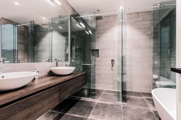 A spacious contemporary bathroom with a double vanity, freestanding vessel basins, a large frameless glass shower enclosure, and a freestanding bathtub partially visible at right. The room features dark wood-look built-in vanity cabinetry, a wood-look benchtop, large grey stone-look wall and floor tiles, extensive mirrors, and chrome fixtures. Bright recessed and linear lighting creates a clean, refined atmosphere.