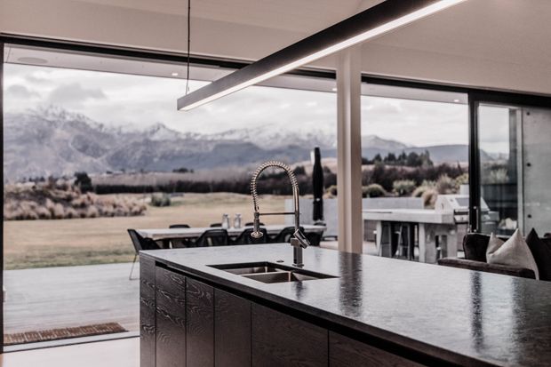 A contemporary open-plan kitchen with a large dark island, integrated sink and high-arc tap, dark textured cabinetry, and a linear suspended pendant light. Full-height glazing opens the room to a broad rural landscape and mountain views, with an adjacent dining and living area visible.