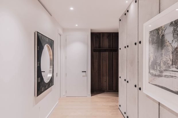 A bright, refined residential hallway or entry corridor with pale timber flooring, white walls and doors, and extensive built-in cabinetry along the right wall. Dark timber wall panelling and open shelving create a contrasting focal point at the end of the corridor, while framed artwork and a round-mirror artwork add visual interest. Recessed ceiling lighting provides even illumination.
