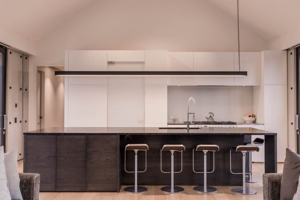 A contemporary open-plan kitchen with a large dark island and integrated breakfast bar seating. The kitchen features extensive white flat-panel cabinetry, a dark stone-like benchtop, a grey tiled or stone splashback, stainless steel tapware, and a long linear pendant light. Glazed doors or windows flank both sides, and partial lounge seating is visible in the foreground. The room has a high pitched ceiling and a restrained monochromatic palette.