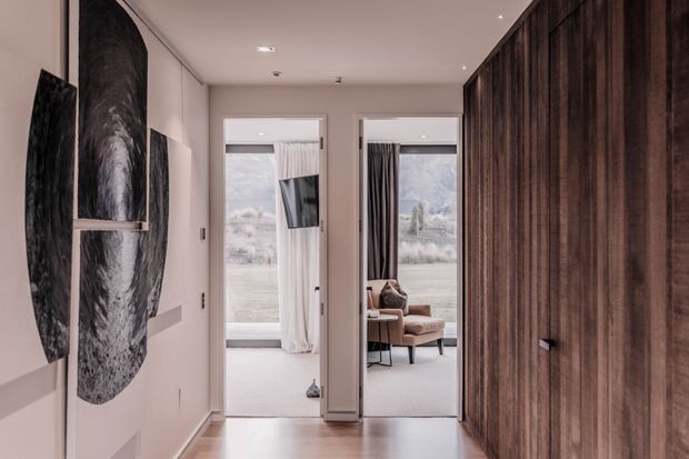 A refined residential hallway with warm timber flooring and a full-height dark timber built-in wall on the right. The left wall features sculptural monochrome artwork, while two glazed openings lead to bright, softly furnished rooms beyond. The neutral palette, concealed lighting, flush detailing, and extensive timber joinery create a calm, contemporary interior.