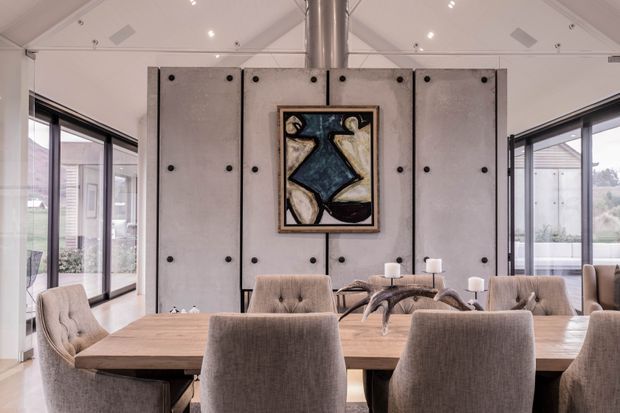 A bright, contemporary dining room centred on a large timber dining table with upholstered dining chairs. The room features expansive black-framed glazing, a tall pale concrete-like feature partition with exposed dark fasteners, a central abstract artwork, a stainless-steel flue or column, and recessed ceiling lighting. Outdoor views are visible through the glazing.