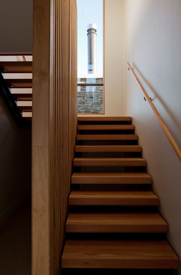 Interior view of a compact modern stairway with warm timber treads, a timber slatted screen wall, dark risers, and a simple timber handrail against a light plaster wall. A tall window brings daylight into the stairwell and reveals a stone-clad exterior wall and cylindrical rooftop element beyond.