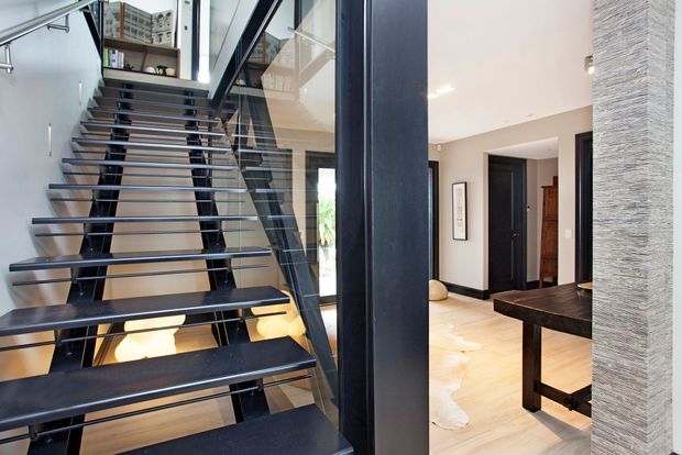 Interior view of a contemporary residential entry and stair hall, featuring an open-riser floating staircase with dark metal stringers and treads, glass balustrade, pale timber flooring, dark interior doors, and a textured vertical feature wall. A dining table is partially visible to the right.