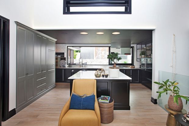 A refined contemporary kitchen viewed from the adjoining living area, featuring a large central island with a light stone benchtop, dark charcoal cabinetry, integrated appliances, a wide horizontal window above the sink, and full-height paneled storage along the left wall. Warm timber flooring and a mustard armchair soften the monochromatic joinery. Recessed ceiling lights and high-level clerestory glazing add brightness and an airy feel.