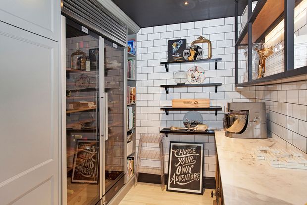 A compact, highly organized walk-in pantry or butler's pantry with floor-to-ceiling glass-front storage on the left, open black shelves against a white subway-tile backsplash, and a light stone countertop on the right. The space includes a stand mixer, decorative servingware, baking and food-storage items, dark ceiling and shelving accents, and warm integrated lighting. The styling is refined and contemporary with a strong black-and-white palette and warm timber details.