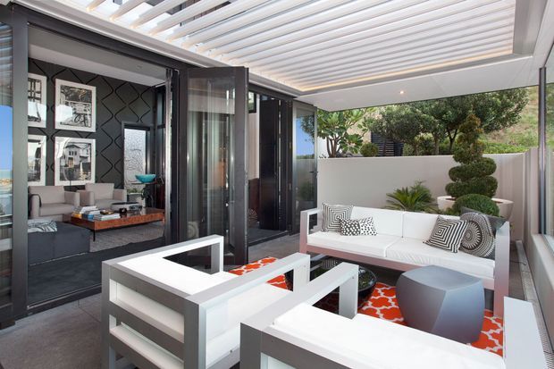 A stylish covered outdoor lounge or terrace adjoining an interior living room through large black-framed folding or sliding glass doors. The space features white outdoor sofas and armchairs, a sculptural grey side table, a bold red-and-white patterned rug, a white wall with clipped topiary and planting, and a translucent slatted roof providing filtered daylight. The adjacent living room has dark patterned wallpaper and framed artwork.