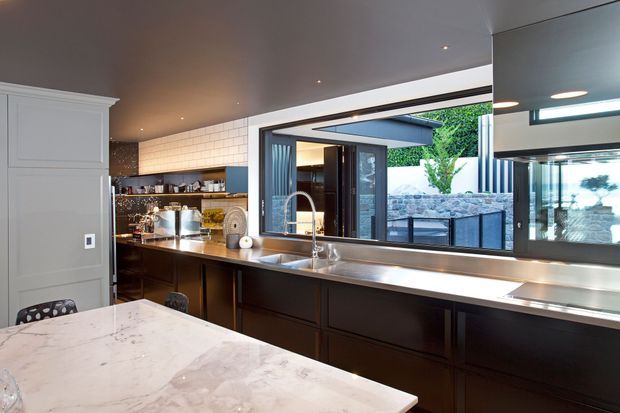 Contemporary kitchen interior with dark timber cabinetry, a long stainless-steel benchtop and integrated sink, a marble island in the foreground, open shelving with glassware, and extensive glazing overlooking a landscaped courtyard. Recessed ceiling lighting and a large mirror or reflective glazing amplify the sense of space.