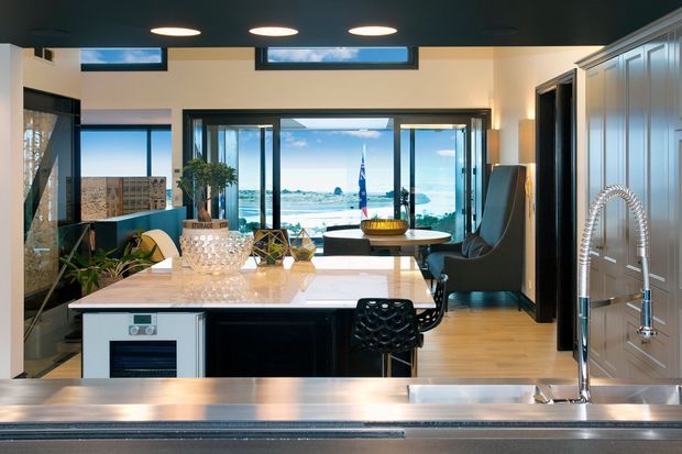A contemporary kitchen viewed from behind the island, with a large white stone island benchtop, dark cabinetry, integrated appliances, and a sculptural black bar stool. Tall glazed doors and windows open the room to a bright coastal outlook, while a dining table and lounge chair sit beyond the kitchen. The space has a dark ceiling with recessed circular lights, light timber flooring, and grey-blue built-in cabinetry along the right wall.