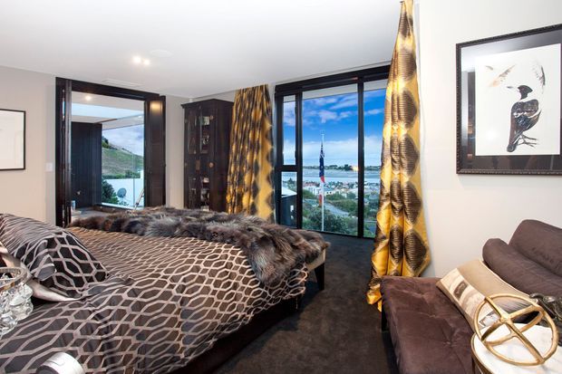 A stylish bedroom with a large upholstered bed dressed in a geometric grey-and-white coverlet and layered faux-fur throws. Floor-to-ceiling black-framed glazing and a glazed door open to a balcony or exterior view, while dramatic gold-and-black patterned curtains frame the windows. A dark wardrobe, framed artwork, sofa, and soft carpet complete the room.