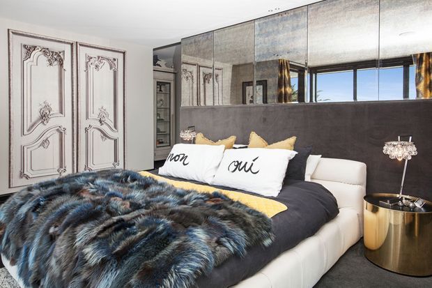 A glamorous bedroom with a large upholstered bed, dark bedding, a dramatic blue-black faux-fur throw, cream and black accent pillows, mirrored wall panels, ornate decorative wardrobe doors, and metallic bedside tables. The room has a strong high-contrast, luxurious aesthetic with reflective surfaces and statement detailing.