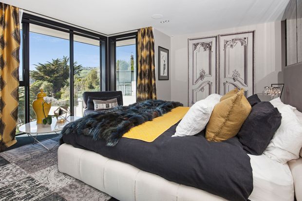 A stylish bedroom with a large upholstered bed dressed in black, white, mustard-yellow, and textured dark throws and cushions. Floor-to-ceiling corner glazing with black frames opens to leafy outdoor views, while ornate decorative double doors, grey wall finishes, patterned curtains, and a patterned rug create a dramatic, eclectic interior.