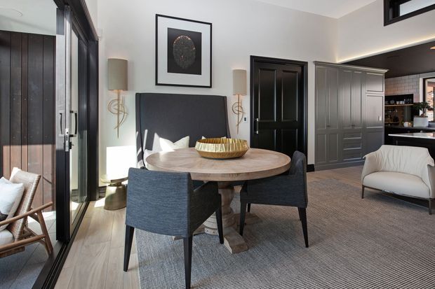 Refined open-plan dining area with a round timber dining table, upholstered dark dining chairs, a black built-in banquette, and a large neutral rug. Black-framed glazed doors and a black feature door create strong contrast, while a dark built-in kitchen or storage area is visible beyond. The room has contemporary-traditional detailing, soft neutral walls, and abundant natural light.