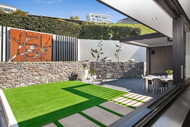 A bright landscaped courtyard viewed from inside through wide-open glazed sliding doors. The compact outdoor space features vivid artificial turf, large rectangular concrete stepping slabs, a rendered boundary wall, a low stacked-stone retaining wall, vertical dark screening, a rust-coloured decorative metal panel, built-in bench seating, and a small outdoor dining setting beneath a covered roof edge. Dense hedge planting screens the rear boundary, with neighbouring modern buildings visible beyond.