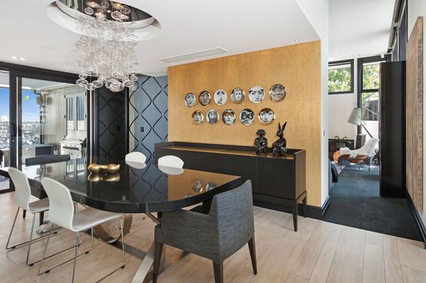 Contemporary open-plan dining area with a glossy black oval dining table, white and upholstered dining chairs, a sculptural glass pendant light, and a black sideboard against a warm timber feature wall. Decorative plates and dark patterned wall panels add visual interest, while a glazed balcony or living area is visible beyond.