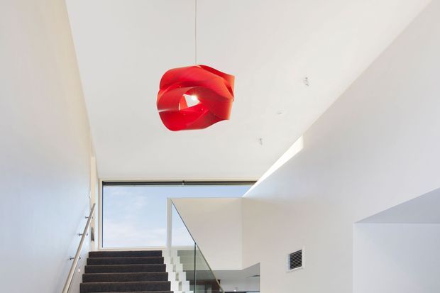 A bright, minimalist interior stairway with dark carpeted steps, white walls and ceilings, a glass balustrade, and a large glazed opening bringing in natural light. A sculptural red pendant light provides a vivid focal accent overhead.