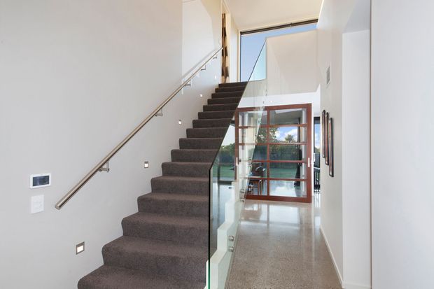 Bright contemporary stairway and hallway with a straight flight of dark carpeted stairs, slim metal handrail, clear glass balustrade, white walls, polished light-toned flooring, and a glazed timber-framed door at the far end. Small recessed wall lights and a high-level skylight or clerestory opening add natural light.