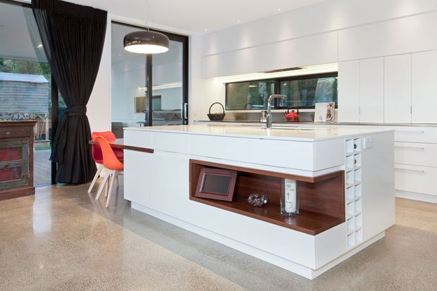 Bright contemporary kitchen dominated by a large white island with integrated sink, polished concrete floor, extensive white flat-panel cabinetry, and a dark timber display niche. A red dining chair and dark curtains add contrast, while a wide horizontal window provides natural light.