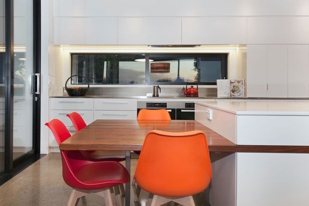 Contemporary open-plan kitchen and dining area with sleek white full-height cabinetry, integrated appliances, a long island with a pale benchtop, and a dark timber dining table. Bright red and orange dining chairs provide bold colour accents. Black-framed glazing and warm under-cabinet lighting add contrast.