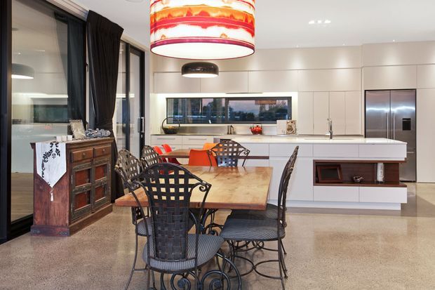 A bright open-plan kitchen and dining area with a large white island, integrated cabinetry, stainless-steel appliances, and a long timber dining table surrounded by ornate black metal chairs. Floor-to-ceiling glazing with dark curtains and a vintage timber sideboard contrast with the streamlined contemporary kitchen. A polished concrete or terrazzo-like floor and statement pendant lights complete the space.