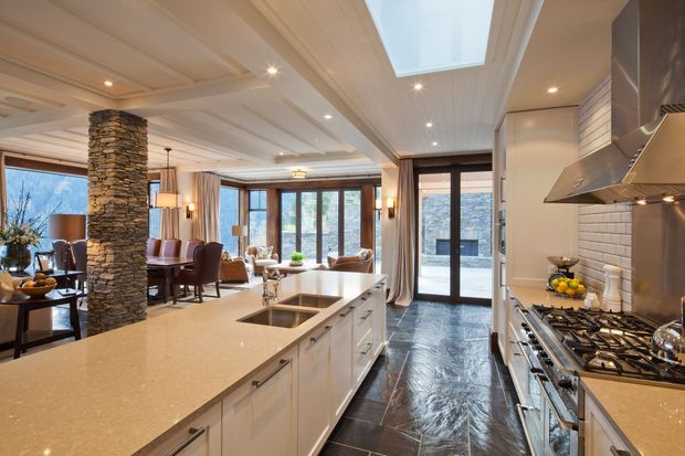 A spacious open-plan kitchen with a large light stone island, white built-in cabinetry, stainless-steel appliances, and a tiled backsplash. The room opens to an adjoining dining and living area with a stone-clad structural column, timber-framed glazing, and a skylight. Dark tiled flooring contrasts with the bright cabinetry and warm timber details.