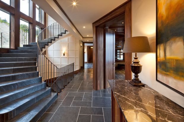A refined residential hallway and stairway with dark stone tiled flooring, a sculptural open-riser staircase, black metal balustrades, tall timber-framed glazing, and rich dark timber wall panelling and door surrounds. A marble-topped console with a table lamp and large abstract artwork add warmth and sophistication.