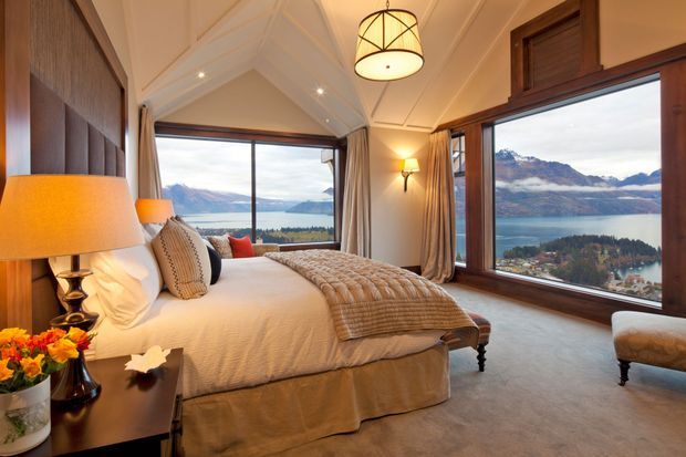 A refined bedroom with a large upholstered bed, layered neutral bedding, bedside lamps, and warm timber-framed windows overlooking a dramatic mountain and lake landscape. The room features a vaulted, panelled ceiling, full-height curtains, dark timber wall panelling, and a soft neutral carpet.