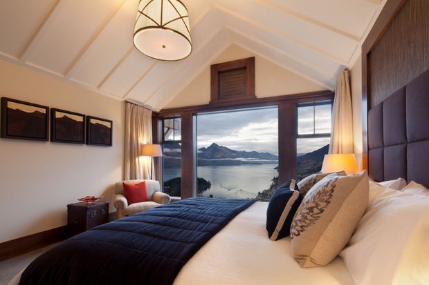 A refined bedroom with a pitched, white-panelled ceiling and large timber-framed glazing overlooking a mountain lake. The room features a king bed with dark quilted bedding, a padded dark upholstered headboard, layered neutral cushions, an armchair, bedside lighting, framed artwork, and warm timber trim.