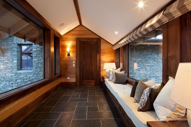 Warm, refined interior corridor or sitting area with dark timber wall panelling and exposed beams, a central timber door, large glazed openings, and grey stacked-stone walls visible through the glazing. A built-in window seat with cushions and table lamps runs along the right side, while large-format dark stone tiles cover the floor. The pitched ceiling and timber detailing create a lodge-like atmosphere.
