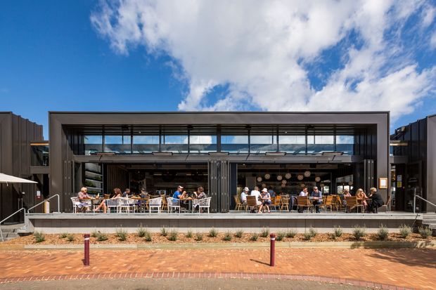 Contemporary two-level commercial hospitality venue viewed from across a brick-paved forecourt. The building has a large rectangular glazed frontage, a deep metal-framed canopy, upper clerestory glazing, dark side volumes, and an open outdoor dining terrace populated with tables and patrons. The architecture is presented against a bright blue sky with landscaped planting along the frontage.