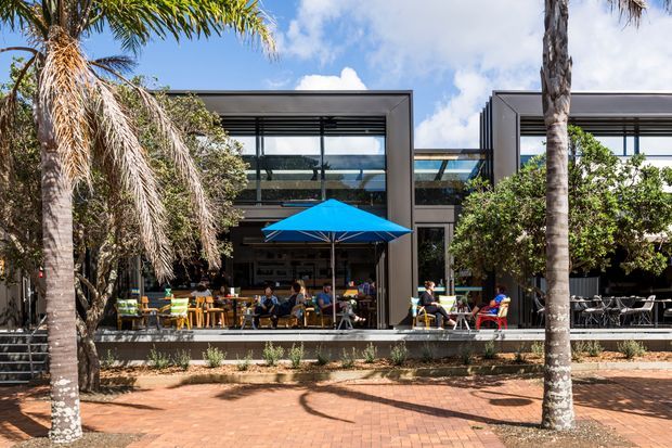 A contemporary commercial café or dining venue viewed from a landscaped pedestrian forecourt. The building has a rectilinear glazed façade with deep dark framing, horizontal louvres, and an open-air terrace furnished with tables and chairs beneath a vivid blue umbrella. Palm trees, brick paving, and abundant greenery create a sunny subtropical streetscape.