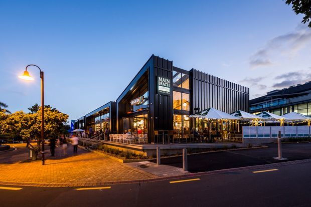 Twilight exterior view of a contemporary multi-level commercial building with a prominent vertical slatted facade, extensive glazing, illuminated restaurant or hospitality spaces, and outdoor dining terraces. The building fronts a paved pedestrian walkway and roadway with street lighting and bollards.