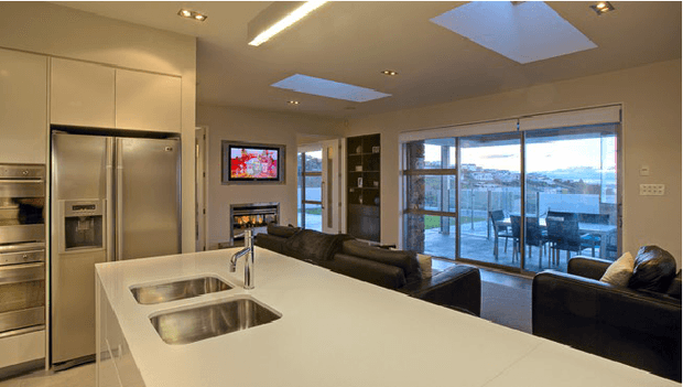 Open-plan contemporary kitchen and living area viewed from behind a large white island bench. The kitchen features stainless-steel appliances, integrated cabinetry, a double-bowl sink and recessed lighting. Beyond the island is a living room with dark sofas, television and extensive sliding glass doors opening to a balcony or terrace, with an expansive urban or coastal view. Multiple skylights bring additional natural light into the space.